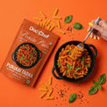 Dodo Chef Protein Pasta Punjabi Tadka with pasta, basil leaves, and a fork on an orange background